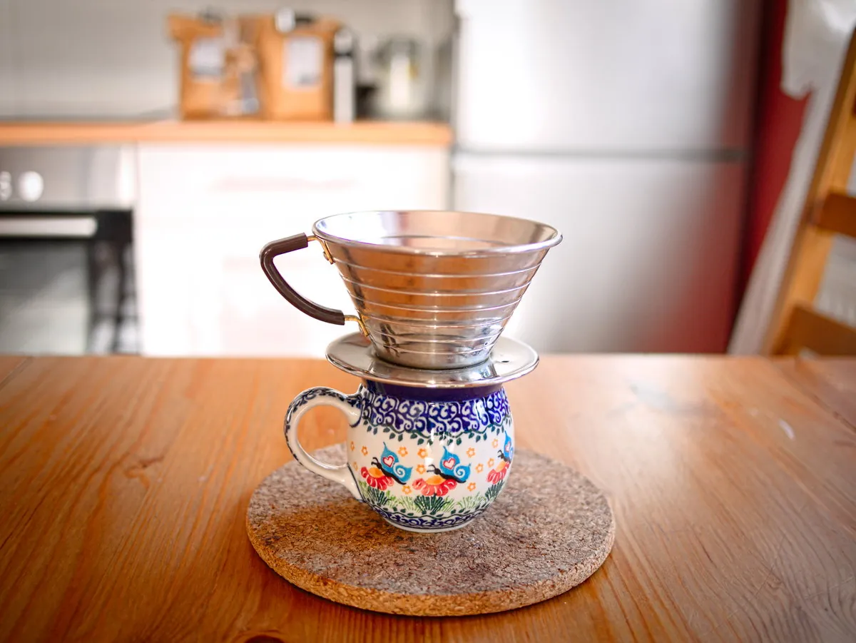 Kalita-Wave-185-Edelstahlhandfilter mit dunklem Griff auf einem gemusterten Keramikbecher.