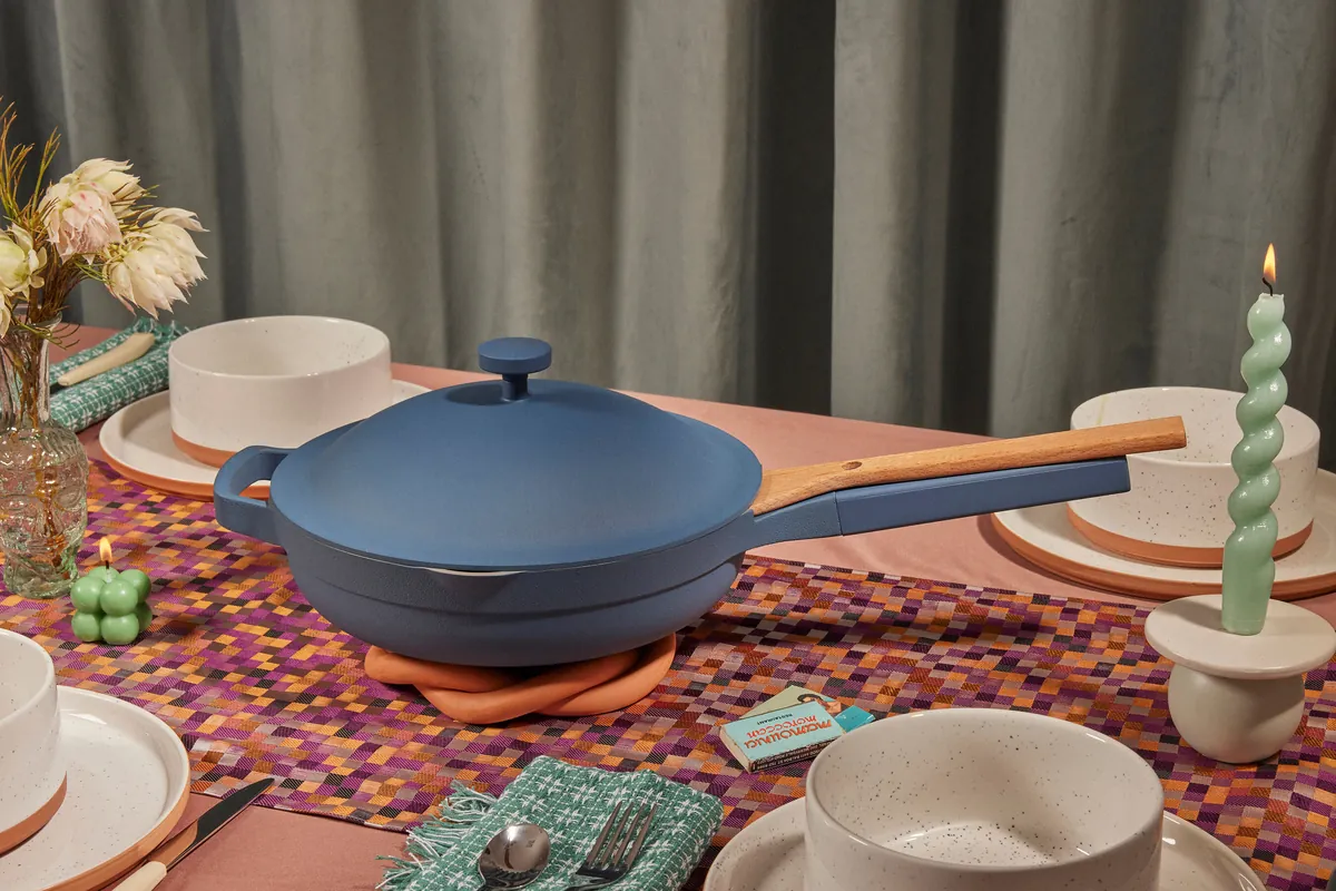 Blue Salt Our Place Always Pan 2.0 with its matching lid and nesting wooden spatula on a dining table.
